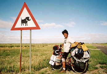ナミビアでの石田さん。看板は、「イボイノシシ注意」の印！世界一周の旅では、各地で出会った仲間とともに走ることもあったとか。